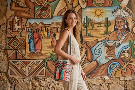 Woman holding a patterned bag in front of a colorful mural with indigenous designs.